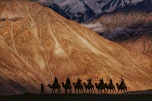 TRAVELERS ON CAMELS ARE SILHOUETTED AGAINST A RANGE OF SANDY MOUNTAINS, THE ONES IN THE BACKGROUND STREAKED WITH SNOW
