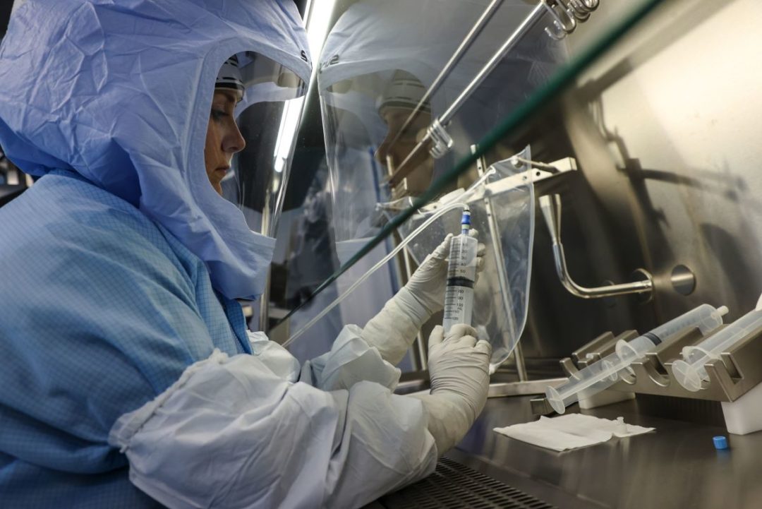 A WORKER IN FULL SAFETY GEAR, INCLUDING HEAD COVERING WITH FACE SHIELD, MANIPULATES A SYRINGE IN A PHARMA FACILITY