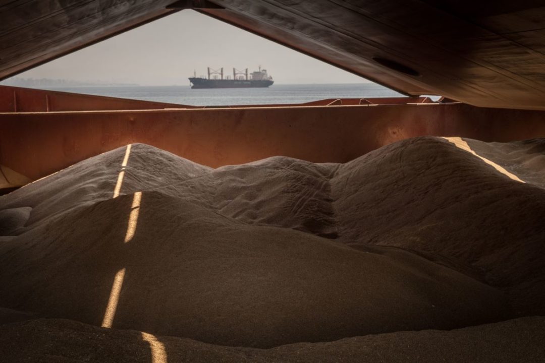A DISTANT SHIP IS SEEN ON THE OCEAN THROUGH A GAP IN THE WALL OF A HUGE GRAIN STORE