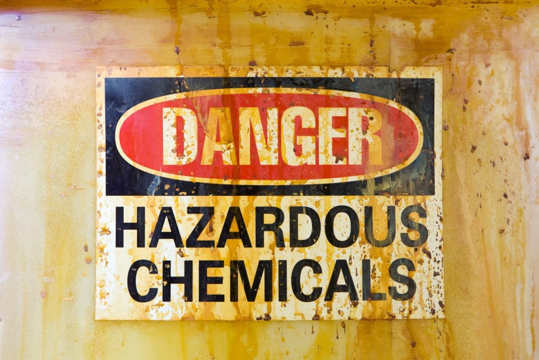 A RUSTED SIGN ON THE SIDE OF A METAL DRUM READS DANGER HAZARDOUS CHEMICALS