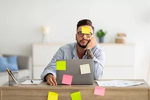 A MAN SITS AT A COMPUTER AT A DESK, COVERED IN POST-IT NOTES, HIS EYES CLOSED IN ANNOYANCE