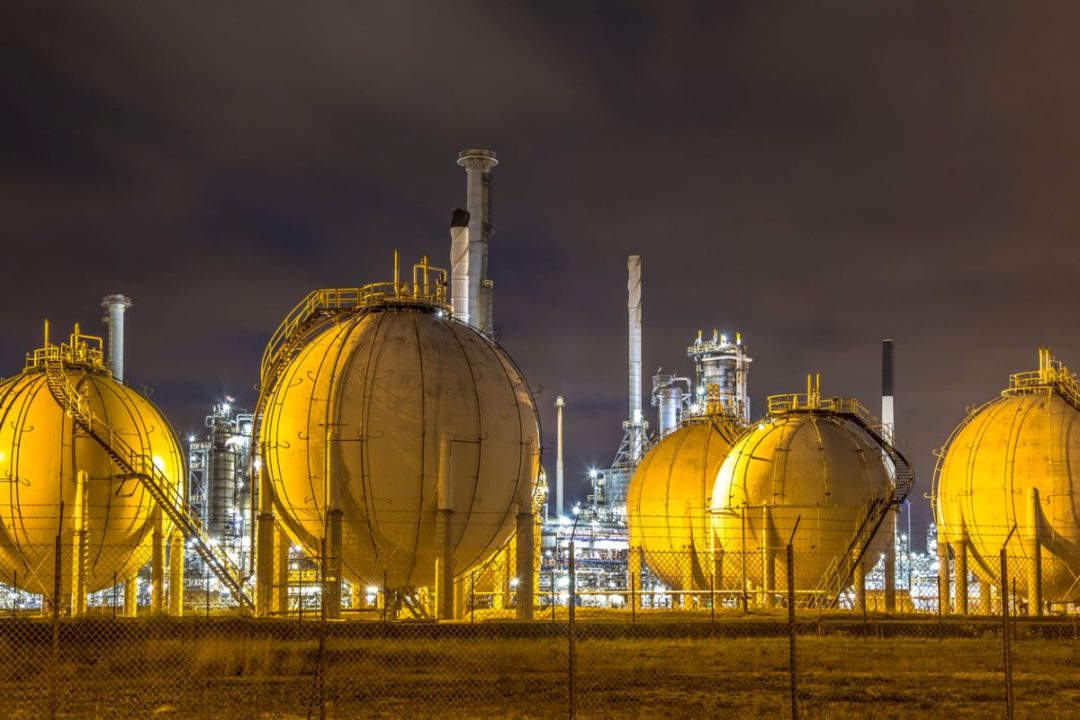 A CLUSTER OF GLOBE-SHAPED LNG STORAGE TANKS SITS UNDER YELLOW LIGHTS AT NIGHT