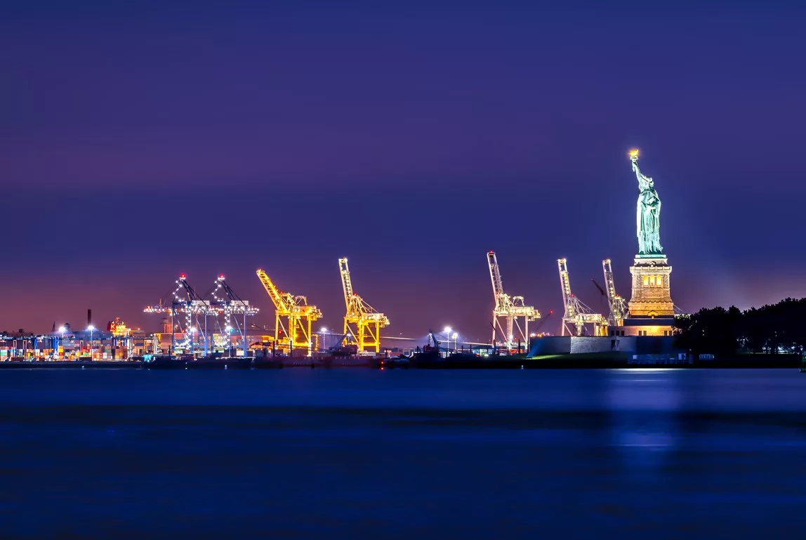 Geceleyin aydınlatılmış Özgürlük Heykeli; arka planda liman vinçleri