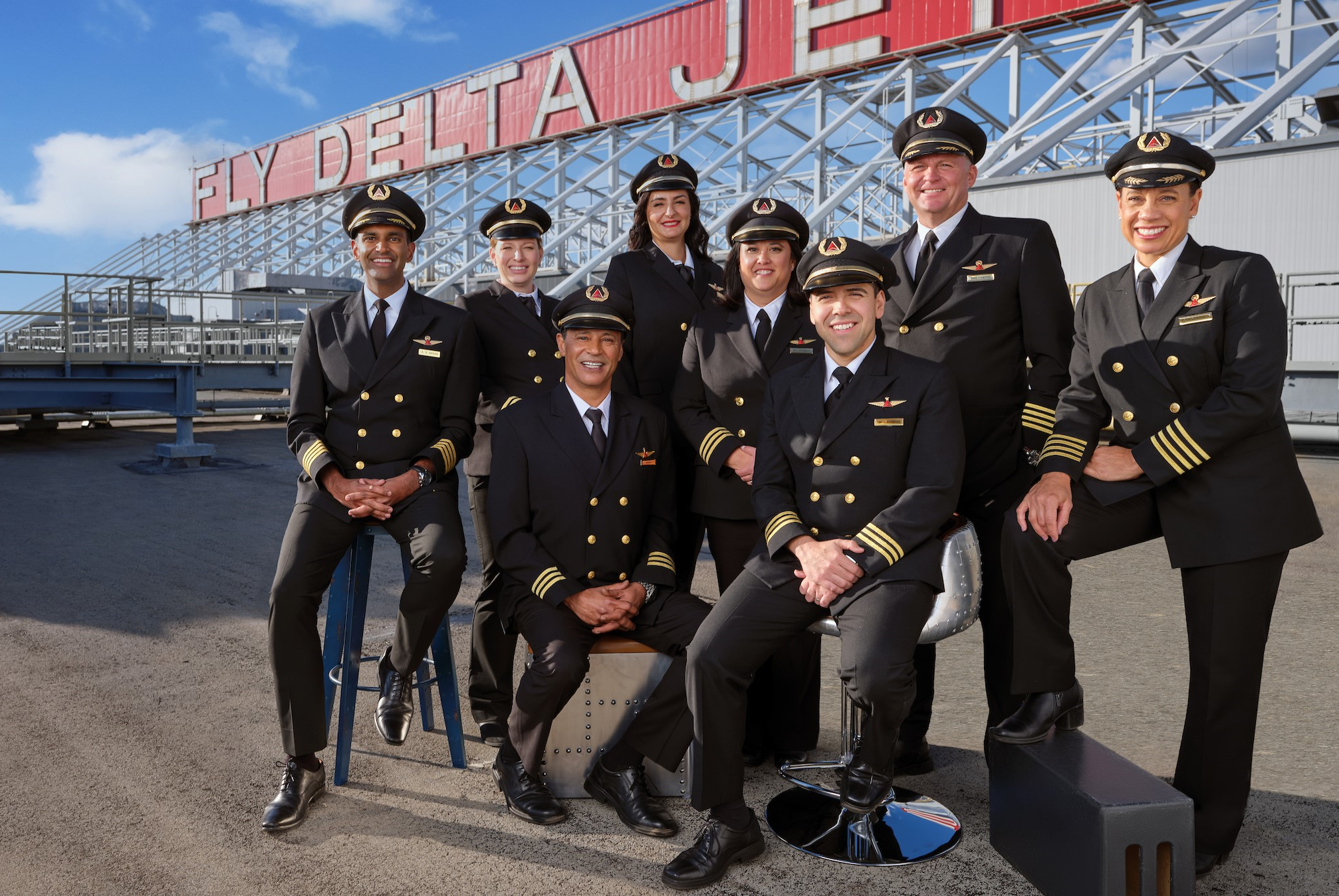 A GROUPING OF MALE AND FEMALE DELTA PILOTS SIT UNDER A SIGN THAT SAYS FLY DELTA JET, ON AN AIRPORT TARMAC