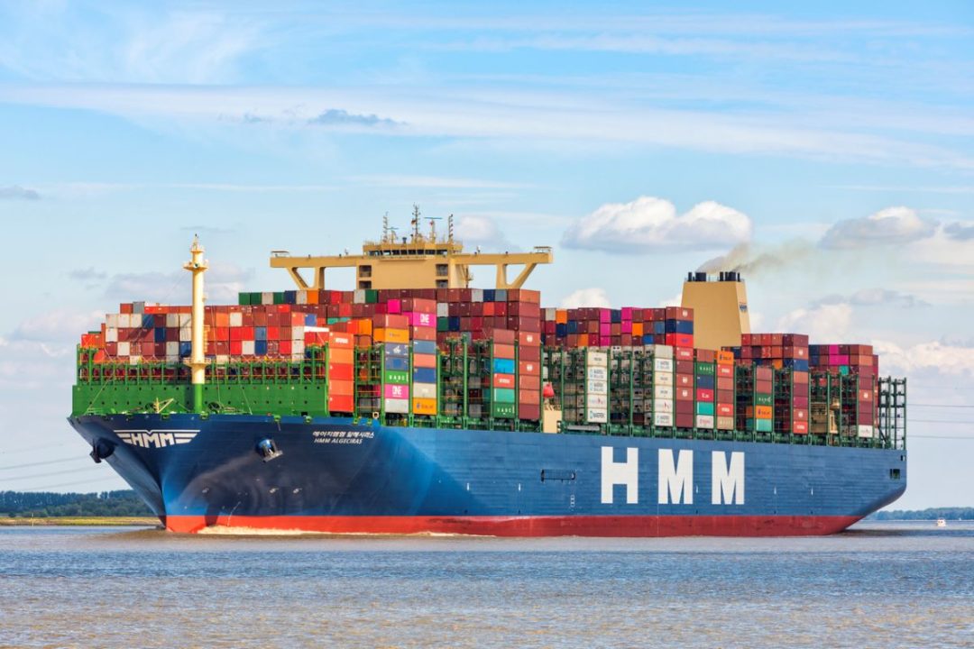 AN ENORMOUS BLUE SHIP, LOADED WITH MULTI-COLORED CONTAINERS, SITS IN RIVER WATERS UNDER A BLUE SKY