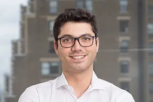 A MAN IN SPECTACLES AND AN OPEN-NECKED WHITE SHIRT SMILES INTO THE CAMERA