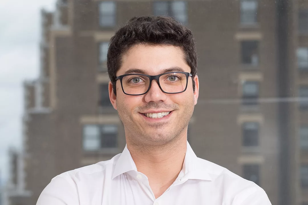 A MAN IN SPECTACLES AND AN OPEN-NECKED WHITE SHIRT SMILES INTO THE CAMERA