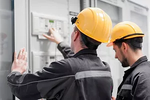 TWO WORKERS IN YELLOW HARD HATS TEND A CONTROL PANEL
