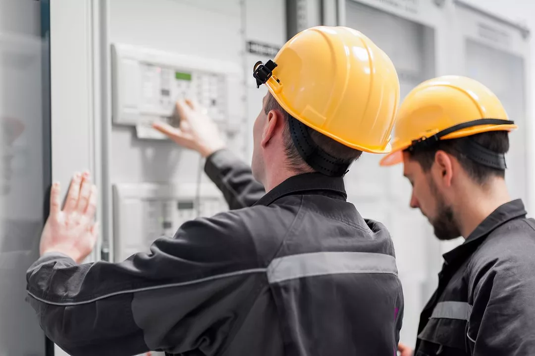 TWO WORKERS IN YELLOW HARD HATS TEND A CONTROL PANEL