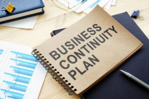 A RING-BOUND FOLDER BEARING THE WORDS BUSINESS CONTINUITY PLAN LIES ON A CLUTTERED DESK