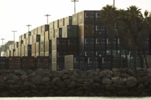 STACKS OF CONTAINERS SIT UNDER TALL LIGHT POLES AND PALM TREES