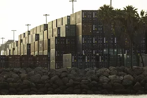 STACKS OF CONTAINERS SIT UNDER TALL LIGHT POLES AND PALM TREES