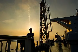 A MAN IN A HARDHAT STANDS ON THE DECK OF A SUPERTANKER, SILHOUETTED BY THE SETTING SUN
