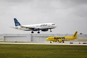 A BLUE AND WHITE JET BLUE PLANE FLIES ABOVE A YELLOW SPIRIT PLANE ON THE TARMAC OF AN AIRPORT