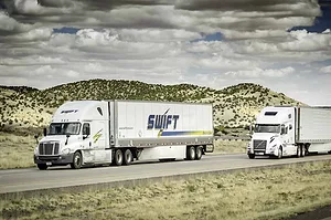 TWO TRUCKS BEARING THE KNIGHT-SWIFT LOGO DRIVE DOWN A HIGHWAY THROUGH DESERT