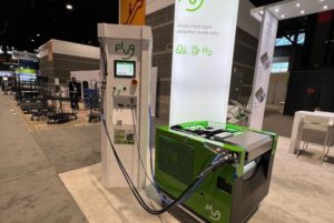 A LARGE GREEN BATTERY SITS HOOKED UP TO A CHARGING STATION ON THE FLOOR OF AN EXHIBITION HALL