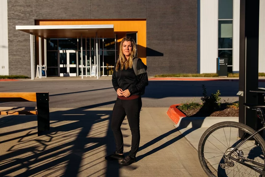 A WOMAN STANDS NEXT TO A BIKE, DAPPLED BY THE SHADOWS OF BICYCLE RACKS.