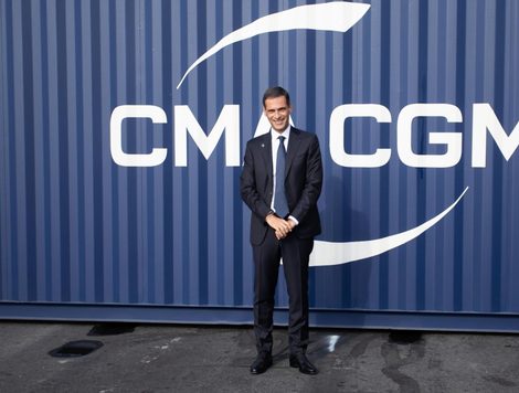 A MAN IN A SUIT AND TIE STANDS SMILING IN FRONT OF A SHIPPING CONTAINER BEARING THE CMA CGM LOGO