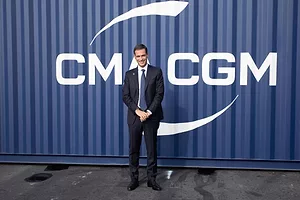 A MAN IN A SUIT AND TIE STANDS SMILING IN FRONT OF A SHIPPING CONTAINER BEARING THE CMA CGM LOGO