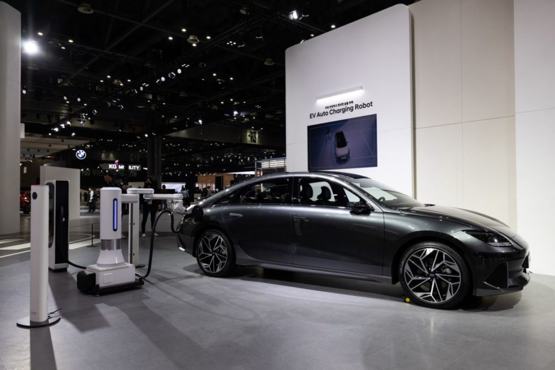A SLEEK BLACK CAR RECEIVES CHARGE FROM AN EV STATION ON A TRADE SHOW BOOTH FLOOR