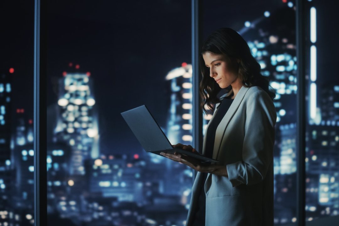 A WOMAN IN A BUSINESS SUIT STANDS TAPPING AT A LAPTOP, THE LIGHTS OF A BIG CITY VISIBLE THROUGH HUGE WINDOWS
