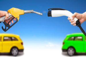 TWO HANDS HOLD DUELING SOURCES OF CAR POWER, ELECTRIC AND GASOLINE, A YELLOW CAR AND A GREEN CAR OUT OF FOCUS IN THE BACKGROUND