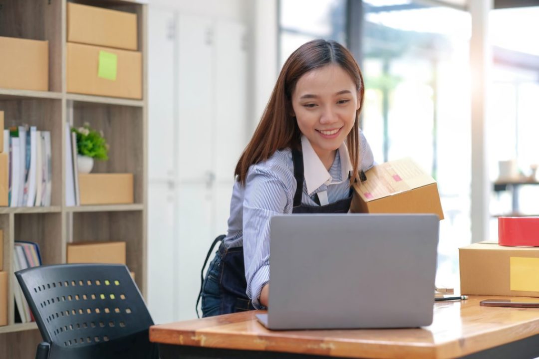A SMILING WOMAN HOLDING A SMALL PACKAGE CONSULTS A LAPTOP IN A SMALL OFFICE