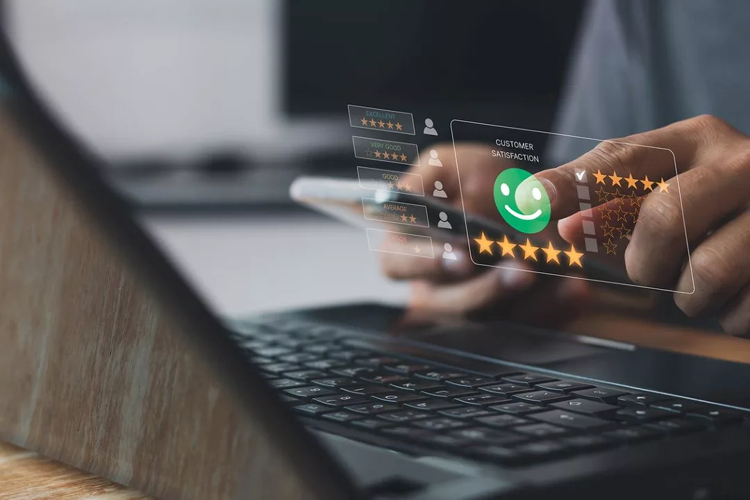 AS A PAIR OF HANDS HOLDS A SMARTPHONE OVER A LAPTOP KEYBOARD, A CUSTOMER SATISFACTION ICON APPEARS