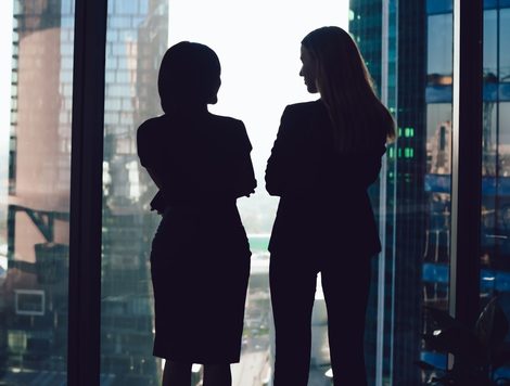 TWO WOMEN TALKING, SILHOUETTED IN A LARGE OFFICE WINDOW, A CITYSCAPE VISIBLE BEYOND
