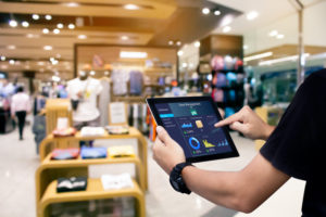 The interior of a retail clothing store can be seen. A manager in a black shirt is using a digital tablet in the foreground of the photo. Photo: iStock.com/B4LLS