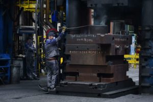 A WORKER IN OVERALLS AND A MASK HANDLES A GIANT STACK OF METAL PARTS
