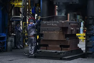A WORKER IN OVERALLS AND A MASK HANDLES A GIANT STACK OF METAL PARTS