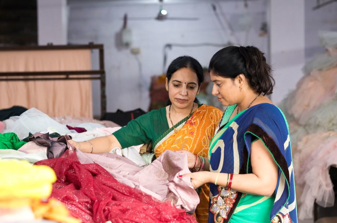 TWO GARMENT FACTORY WORKERS WEARING SARIS HOLD A STACK OF CLOTH PIECES