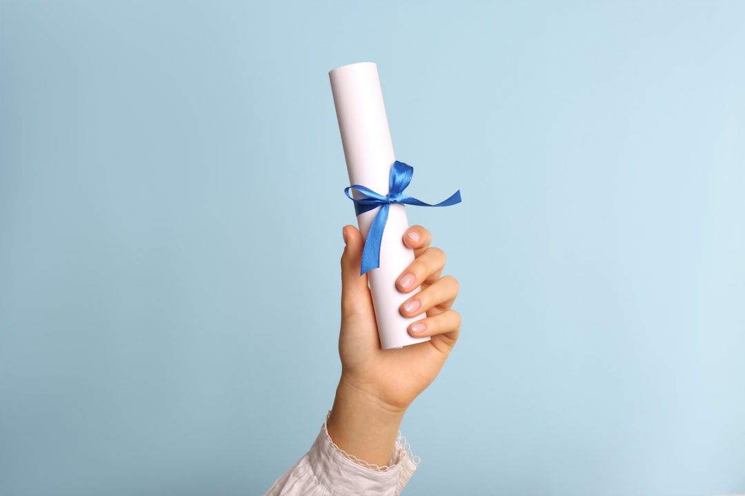 A person's hand can be seen holding a rolled diploma with a ribbon on it in front of a light blue background. Photo: iStock.com/Liudmila Chernetska