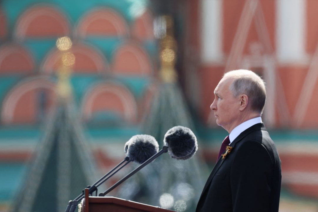 VLADIMIR PUTIN STANDS AT A MICROPHONE, THE KREMLIN OUT OF FOCUS IN THE BACKGROUND
