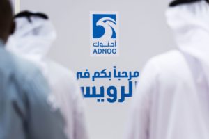 TWO MEN IN WHITE DISHDASHA ROBES AND GUTHRAIN HEAD-DRESSES REGARD A BLUE AND WHITE SIGN FOR ADNOC ON A WALL