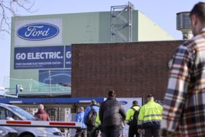 PEOPLE MILL OUTSIDE A BUILDING THAT HAS WRITTEN ON THE SIDE IN LARGE LETTERS: FORD GO ELECTRIC