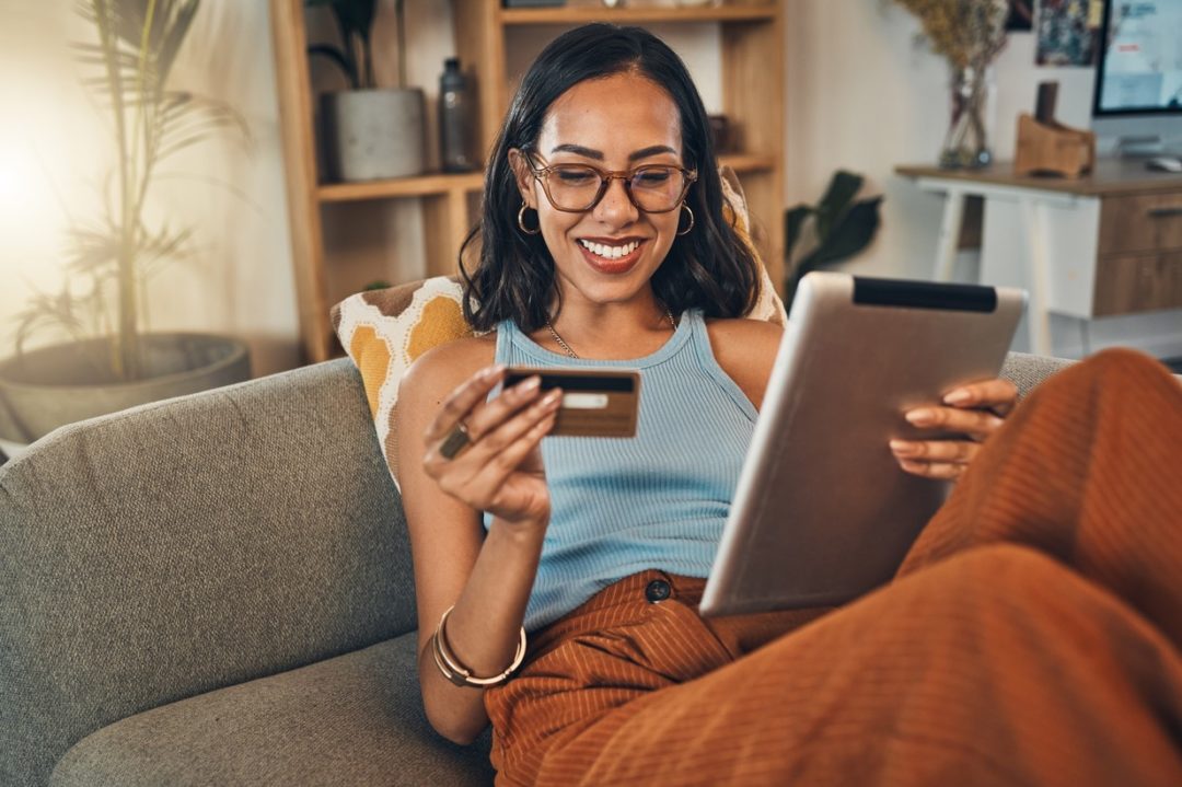 A SMILING WOMAN SITS ON A SOFA, HOLDING A COMPUTER TABLET IN ONE HAND AND A CREDIT CARD IN THE OTHER