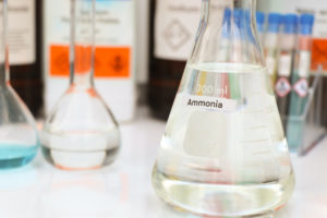 A GLASS BEAKER SURROUNDED BY OTHER CONTAINERS SITS ON A DESK IN A LABORATORY WITH A STICKER ON THE FRONT THAT READS "AMMONIA"