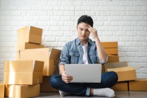  A MAN SITTING IN A PILE OF BOXES, HOLDING A LAPTOP, LOOKS DISTRESSED