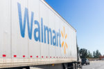 A LARGE WHITE WALMART TRACTOR TRAILER IS DRIVING ON A FREEWAY BEHIND A PICK-UP TRUCK ONE LANE OVER.