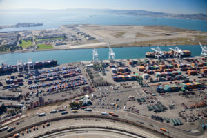 TWO OCEAN CARRIERS ARE DOCKED AT THE PORT OF OAKLAND ON A SUNNY DAY ADJACENT TO A PARKING LOT AND BUSY HIGHWAY.