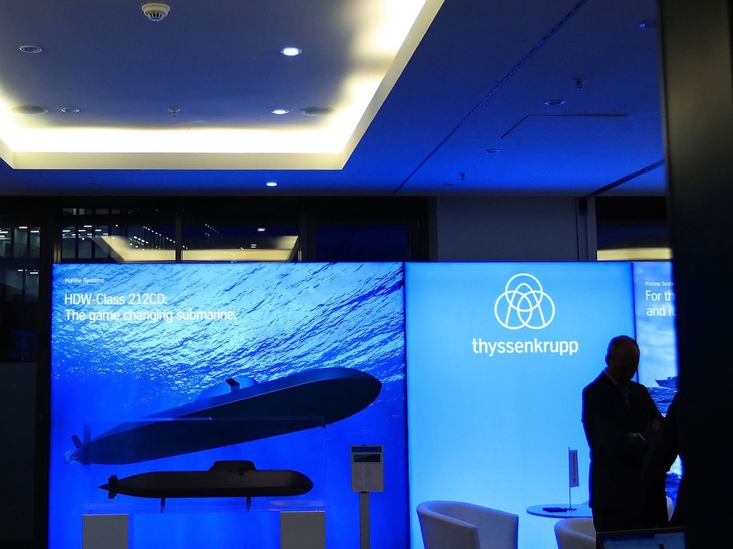 A SUBMARINE EXHIBIT PUT ON BY THYSSENKRUPP SHOWS SUBMARINES IN THE OCEAN ON THE LEFT SIDE OF A SCREEN AND THE COMPANY LOGO ON THE RIGHT SIDE. 