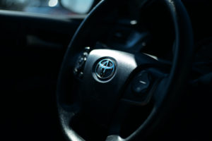 THE BLACK INTERIOR OF A CAR SHOWS A BLACK STEERING WHEEL WITH A TOYOTA LOGO IN THE CENTER OF IT.