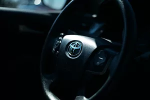 THE BLACK INTERIOR OF A CAR SHOWS A BLACK STEERING WHEEL WITH A TOYOTA LOGO IN THE CENTER OF IT.