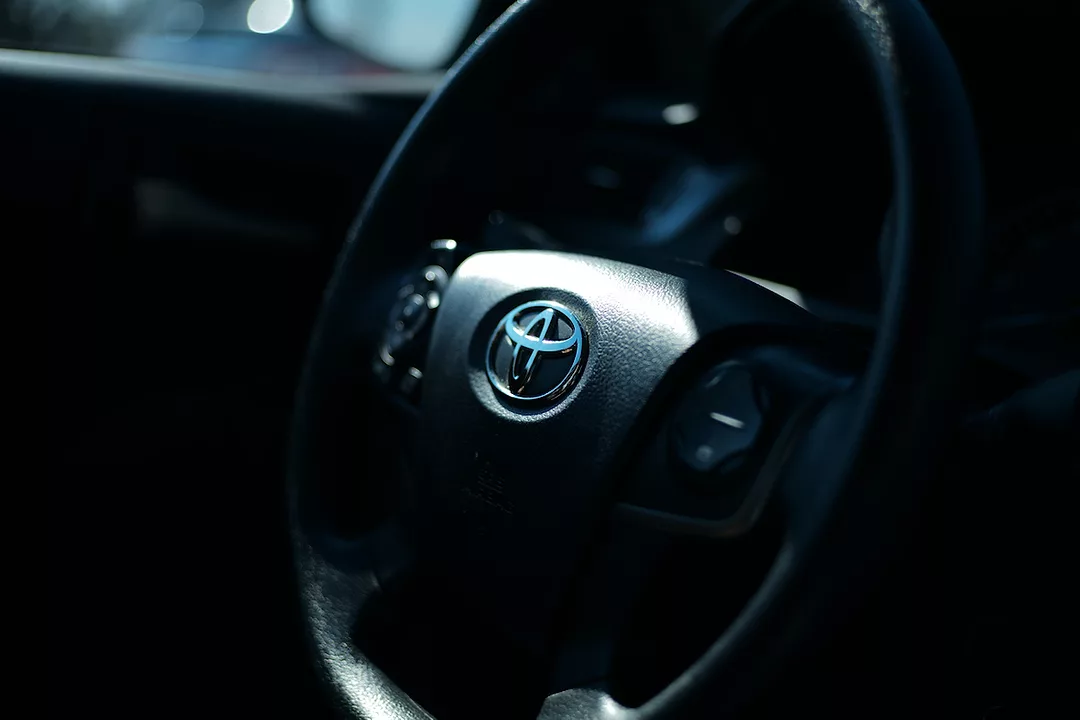 THE BLACK INTERIOR OF A CAR SHOWS A BLACK STEERING WHEEL WITH A TOYOTA LOGO IN THE CENTER OF IT.