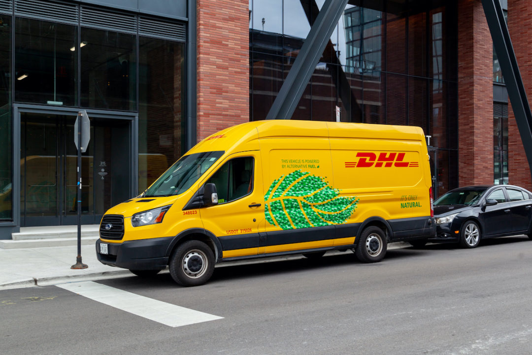 A DHL TRUCK IS PARKED ON THE STREET IN FRONT OF A STOP SIGN OUTSIDE OF AN OFFICE BUILDING WITH A BLACK CAR PARKED BEHIND IT.