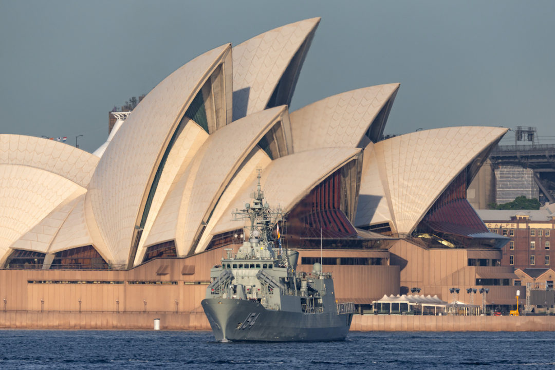 A ROYAL AUSTRALIAN NAVY SHIP CAN BE SEEN FLOATING ON THE WATER WITH THE SYDNEY OPERA HOUSE BEHIND IT.