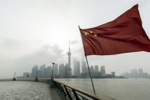 A CHINESE FLAG IS BLOWING IN THE WIND ON A CLOUDY DAY WITH THE CITY OF SHANGHAI IN THE BACKGROUND.