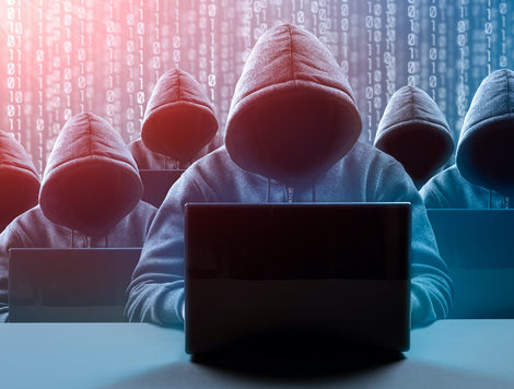 SEVEN HOODED FIGURES, WHOSE FACES CANNOT BE SEEN, ARE SITTING IN FRONT OF LAPTOPS WITH DIGITAL BINARY IN THE BACKGROUND.